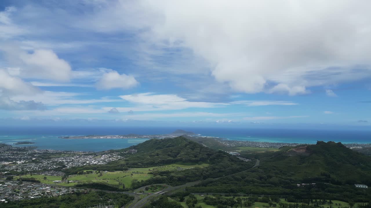 High-altitude drone rotating over Big Island, showing city, mountains, coastline, and cloudy sky.