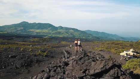 Drone-aerial-flying-over-two-travelers-standing-on-a-rock,-looking-at-a-rocky-landscape-in-Pacaya,-Guatemala