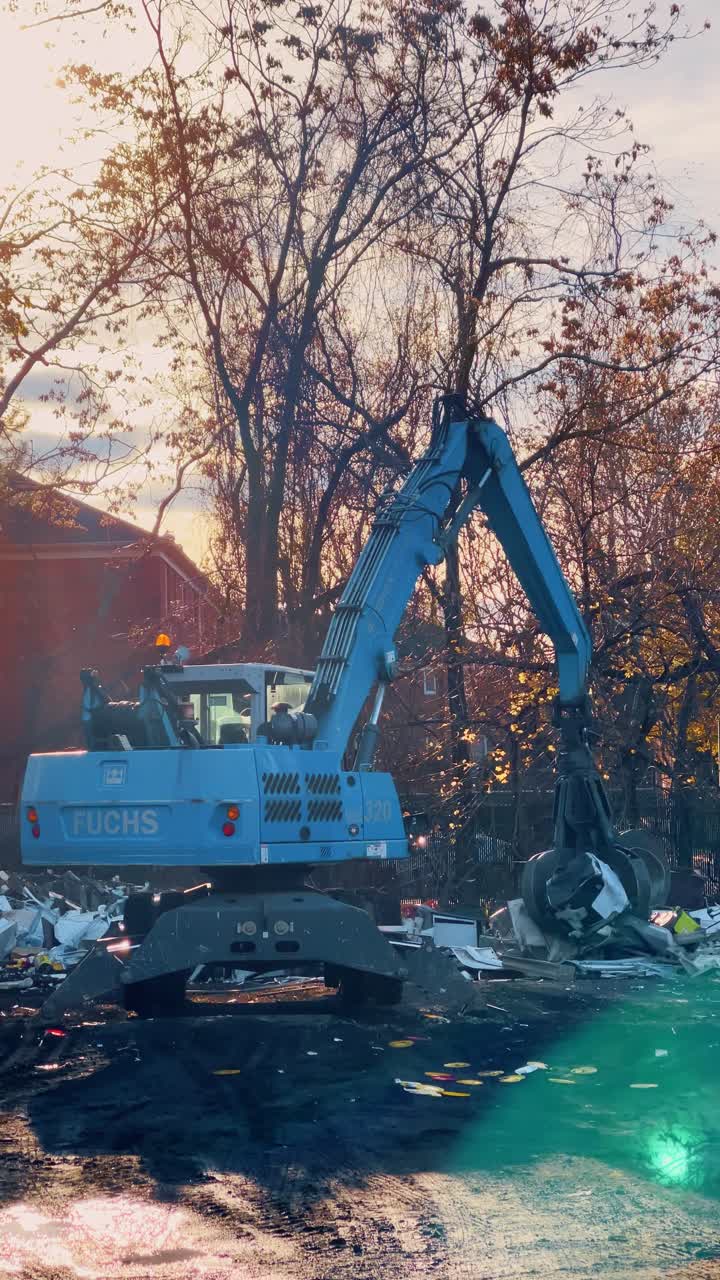 New York, USA, 20 ottobre 2025: Gru industriale per movimentazione materiali che carica rottami metallici presso uno sfasciacarrozze. Escavatore idraulico blu con benna mordente che sposta detriti metallici e materiali da riciclo all'aperto vicino agli alberi.