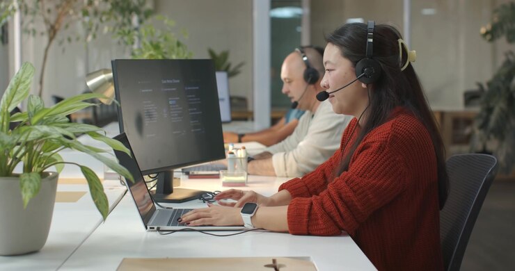 Diverse coworkers with Asian woman in red sweater taking call, typing on laptop at shared desk