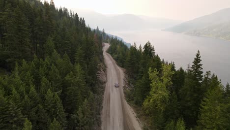 Silver-car-driving-along-a-dusty-forest-service-road-during-wildfire-season-with-Adams-Lake-in-the-background