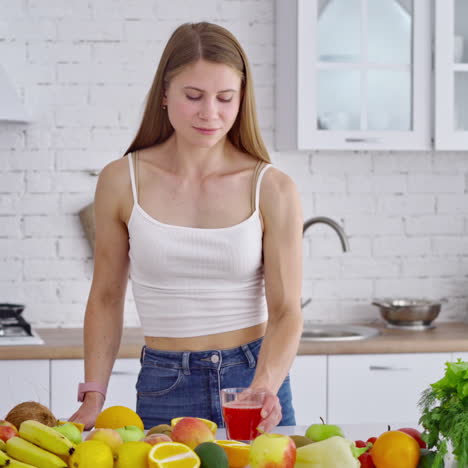 young woman drinking fresh juice. beautiful lady with glass of tasty juice standing near the table with organic fruit in the modern kitchen. losing weight concept.