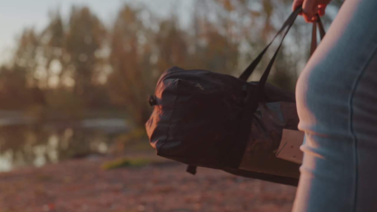 Primo piano di donna che trasporta un borsone da campeggio in riva al lago durante un tramonto dorato, mani che stringono le cinghie, gambe vestite di jeans che camminano su una riva di ciottoli, borsa appoggiata a terra tra foglie autunnali, preparazione tranquilla.
