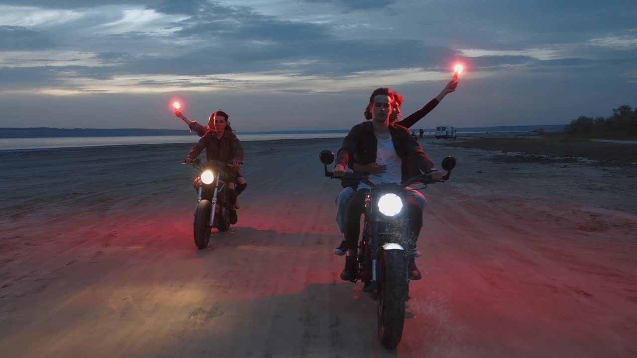 dos parejas jóvenes montando motocicletas antiguas con bengalas de señal rojas ardiendo después de la puesta de sol en la playa, en cámara lenta