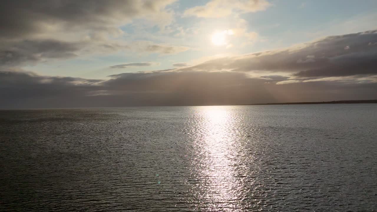 Still camera captures the sun setting over tranquil ocean waters, with dramatic clouds and golden reflections, creating a peaceful, atmospheric mood