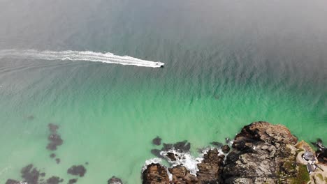 Toma-Aérea-De-Una-Lancha-Rápida-Que-Navega-En-Aguas-Turquesas-Y-Pasa-Por-Rocas-De-Cabecera-En-St.-Ives,-Cornualles,-Inglaterra