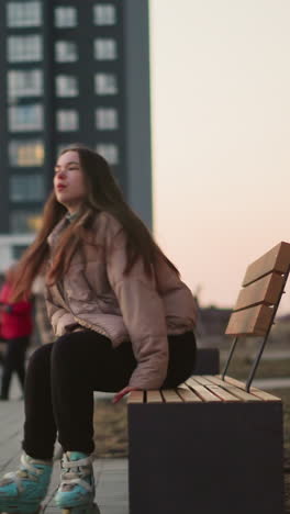 a girl in a peach jacket and black trousers is seen rollerblading towards a park bench in an urban environment. as she sit and rest, other people can be seen in the background