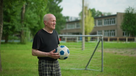 an elderly man rolls a soccer ball in his hand with a focused expression, standing in a grassy field with a building and goalpost in the background