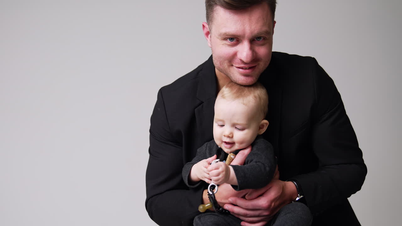 Caucasian man wearing black jacket holding a little cute baby in hands. Little kid is focused on the pacifier. Dad and son concept. White backdrop.