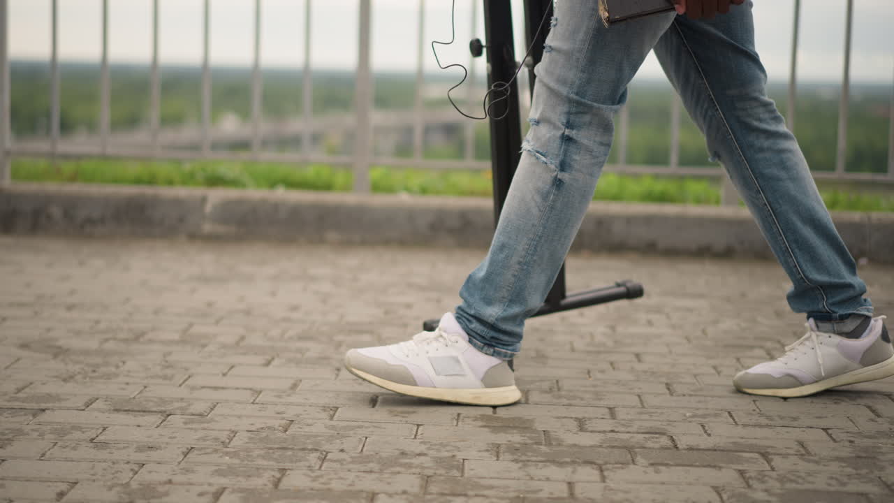 Gatemusikant går med stativ, dongeribukse og hvite joggesko på fortauet, bærer mikrofonstativ mellom settene, overskyet byrekkverk i bakgrunnen, sakte gange som fanger avreise etter utendørskonsert, stemningsfullt.