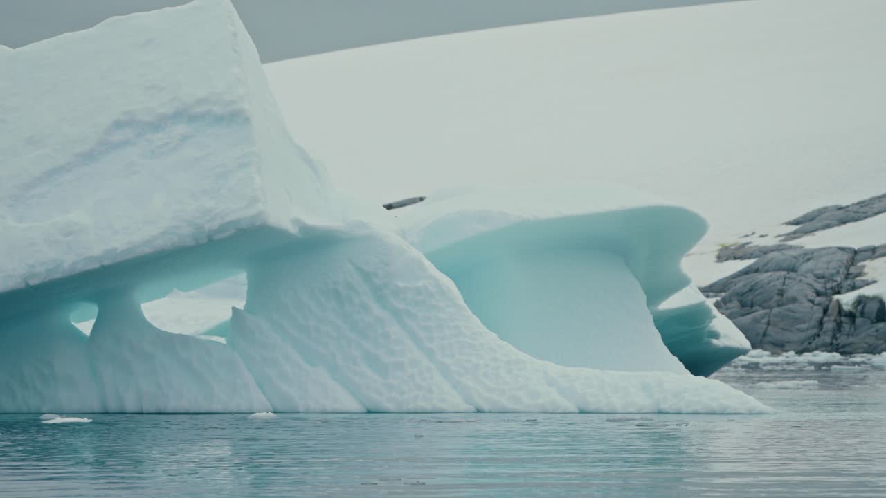 Low-angle tracking shot reveals wave-carved arches and glowing turquoise caverns in massive sculpted iceberg floating near rocky Antarctic coastline