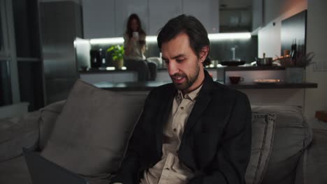 A-happy-brunette-man-with-stubble-in-a-black-suit-and-beige-shorts-works-on-a-Gray-laptop-while-sitting-on-a-sofa-in-a-modern-apartment-near-his-girlfriend-who-is-sitting-in-the-kitchen-and-drinking-tea