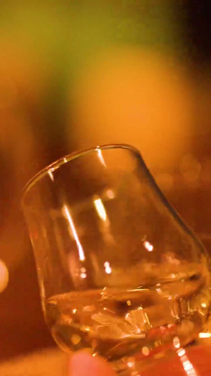 Una mano fa roteare un bicchiere di whisky su un tavolo, il liquido dorato che cattura la luce soffusa e calda dell'ambiente di un bar