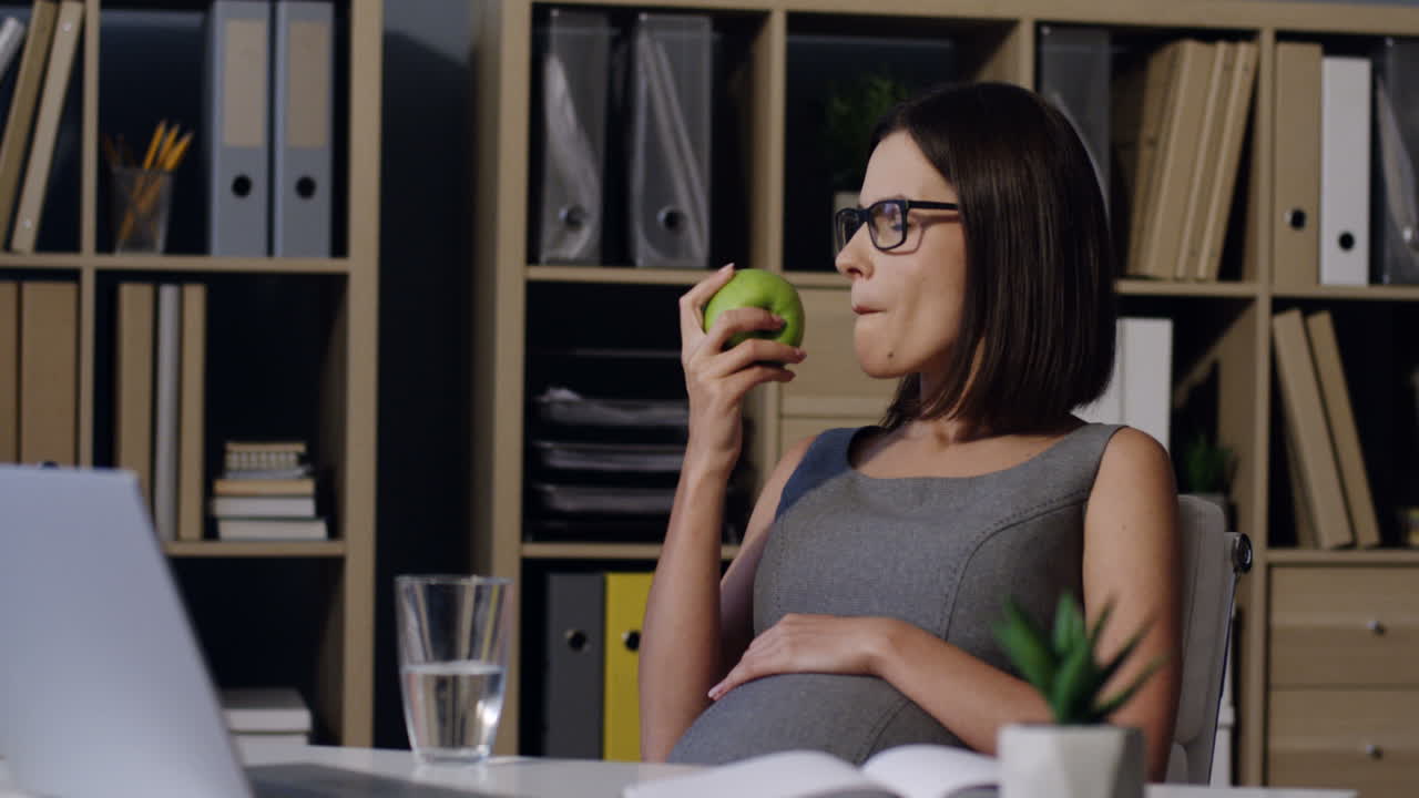 Pregnant Woman In Glasses Biting A Green Apple While Resting In The Office At Her Working Place