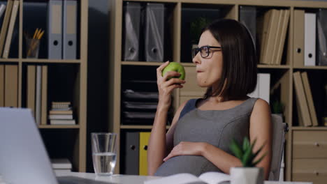 pregnant woman in glasses biting a green apple while resting in the office at her working place