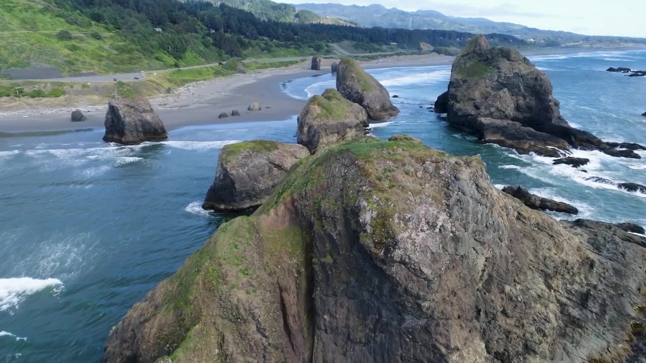 Un filmato cinematografico con drone scivola in avanti sopra i faraglioni lungo la spiaggia di Ariya, rivelando imponenti faraglioni, le onde del Pacifico e una costa frastagliata vicino a Branson Carlton's View sulla costa meridionale dell'Oregon