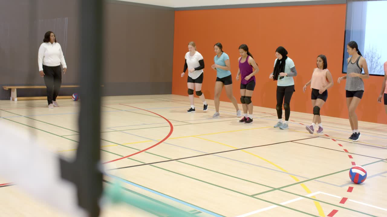 Allenatrice afroamericana che entra da sinistra, allena la squadra che esegue affondi in palestra con una palla da pallavolo.