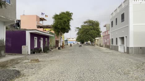 a tranquil cobblestone street in sal rei, cape verde, lined with vibrant, colorful buildings and lush trees. the peaceful atmosphere highlights the charm of this picturesque village scene