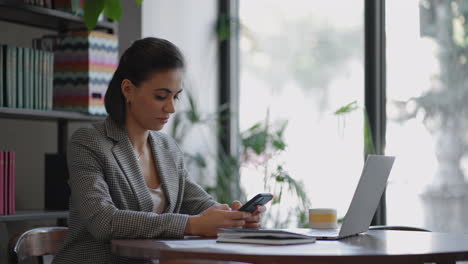 Stylish-young-woman-uses-a-smartphone-surf-the-internet-sitting-at-a-table-in-modern-cafe-smiling.-young-business-woman-indoors-in-office-using-mobile-phone