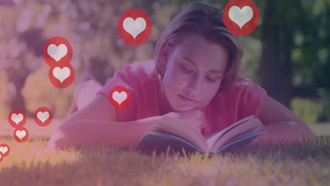 animation of heart icons over caucasian woman lying on lawn and relaxing with book