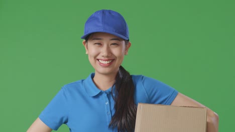 푸른 유니폼을 입은 아시아 여성 택배사가 에 손을 고 미소를 지으며 스튜디오에서 녹색 스크린 배경에 박스를 배달합니다.