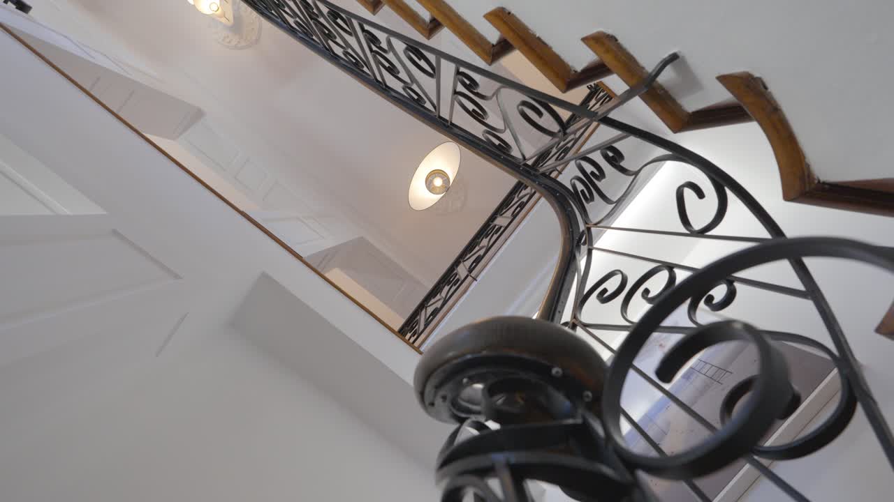 escalera en cámara lenta, con su barandilla de hierro forjado y sus paredes blancas con molduras, los escalones están hechos de madera