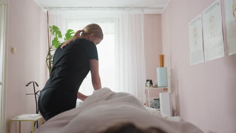 massage therapist in black outfit seen from back view massaging client foot in softly lit therapy room with anatomical posters on wall, bright curtains, and indoor plant adding to tranquil setting
