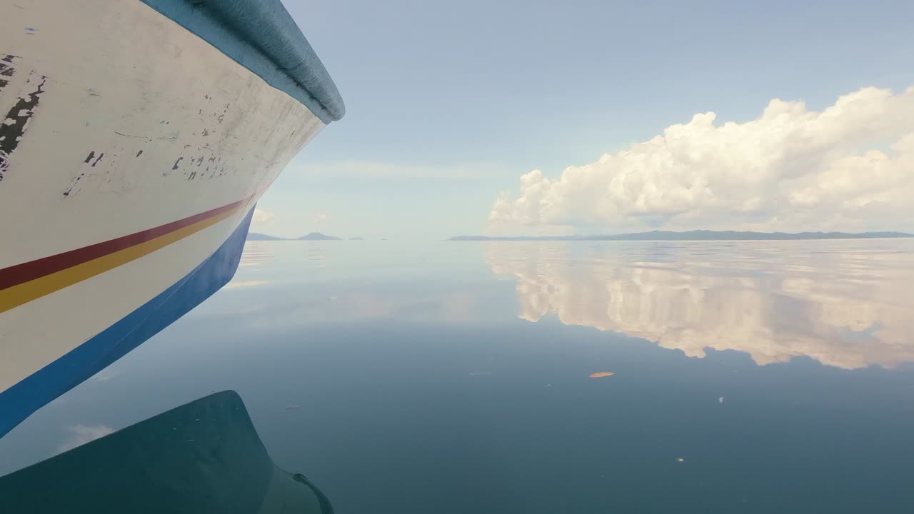 Serene shot of a boat gliding over a mirror-like sea, with clouds and distant islands reflected on the still water. The calmness of the scene and the vast ocean evoke solitude in a tropical paradise.