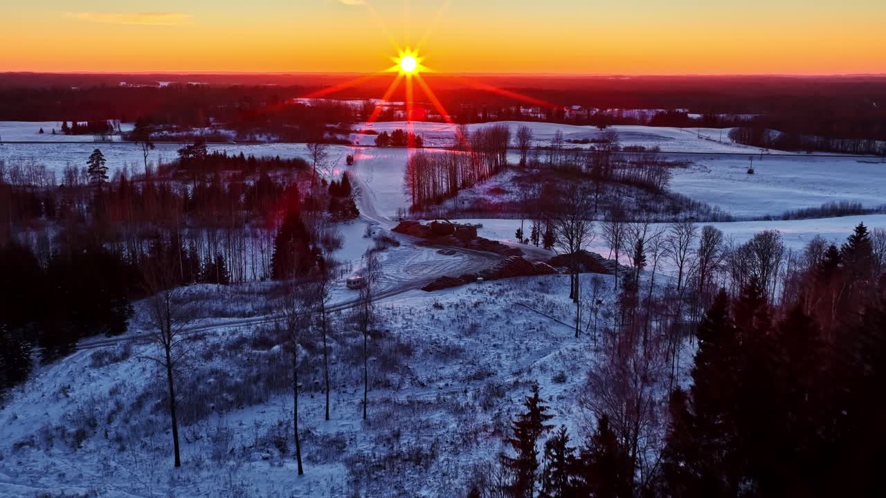 Talvinen auringonlasku lumisen maiseman yllä puiden ja etäisten peltojen kanssa.