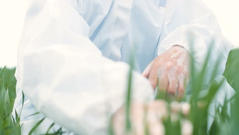 close-up view of caucasian researcher man in protective suit and goggles sitting on green field and touching organic herb of wheat