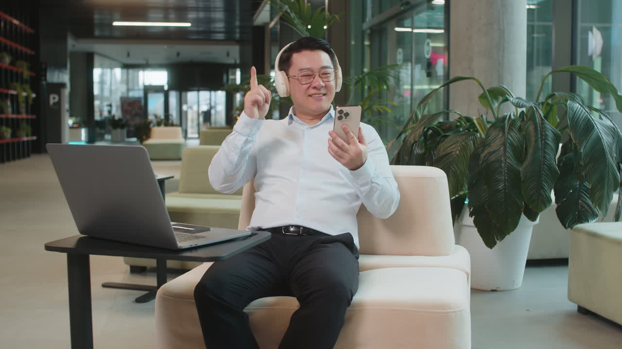 Excited mature businessman in headphones listening energetic music on smartphone in office lobby