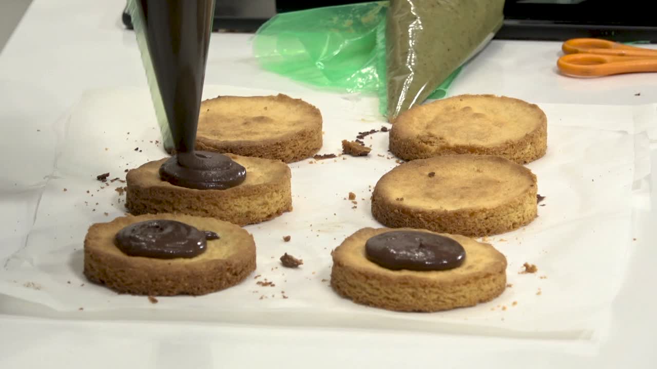 Participants are engaged in a hands-on culinary class, skillfully piping chocolate filling onto freshly baked cookie bases on a workspace. The atmosphere is lively and educational.