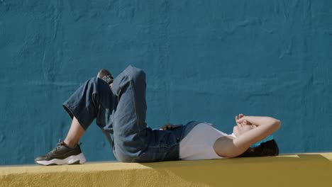 young woman smiles listen to music with wireless headphones while laying down wearing overalls during a sunny day with a yellow and blue wall background