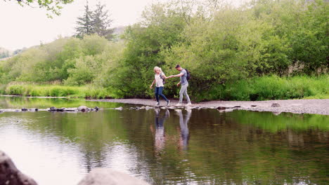 Slow-Motion-Shot-Of-Young-Couple-Using-Stepping-Stones-To-Cross-River-Whilst-Hiking-In-UK-Lake-District