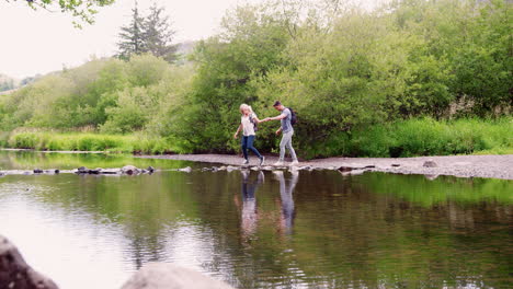 slow motion shot of young couple using stepping stones to cross river whilst hiking in uk lake district