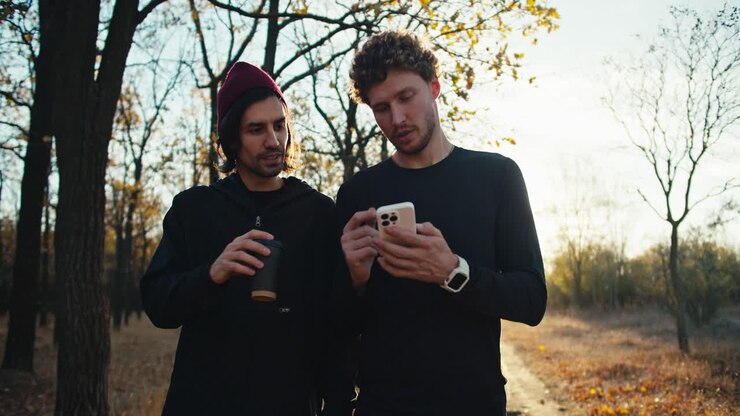 A confident and happy man with curly hair in a black sports uniform with a white phone in his hands walks and communicates with his friend a man with a red during their walk in the autumn forest in the evening