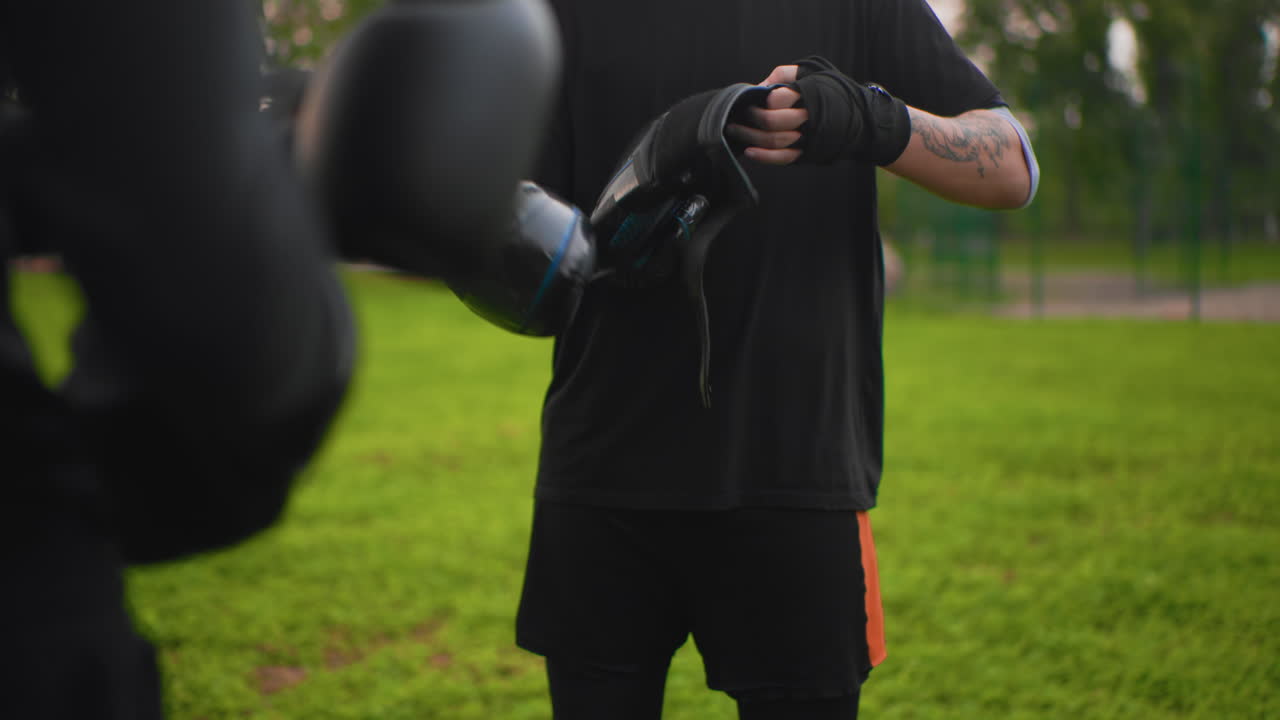 white man tightening boxing gloves outdoors, tattooed forearms, closeup midsection, preparing for sparring partner, focused breathing, black training shirt, green park background, dusk light, casual