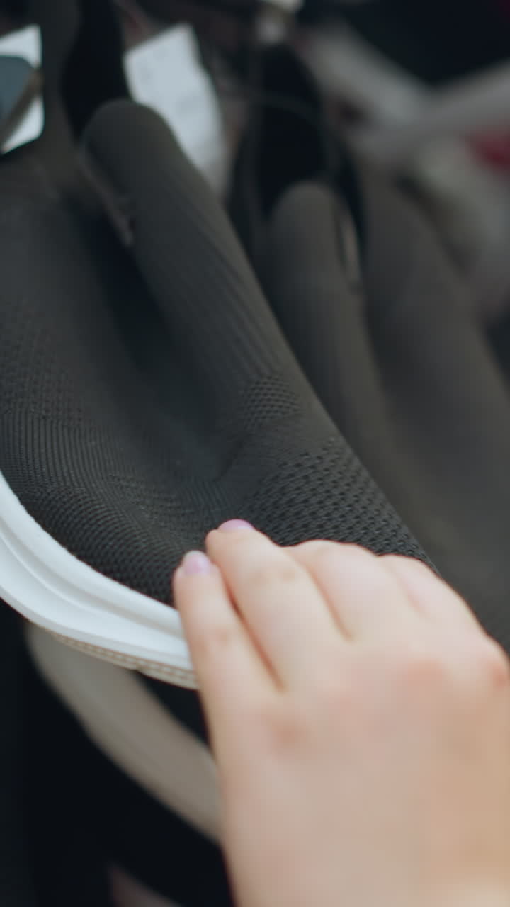 신발 가게에서 분홍색 드레스를 입은 여자의 손은 두 손으로 검은 스니커 발바닥을 면밀히 조사하고, 다양한 신발로 가득 찬 선반은 배경에서 약간 흐릿합니다.