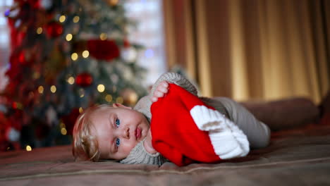 lovely sweet baby boy with fair hair lies on the sofa. cute child holds a santa cap and pulls it to his mouth. blurred christmas tree at backdrop.