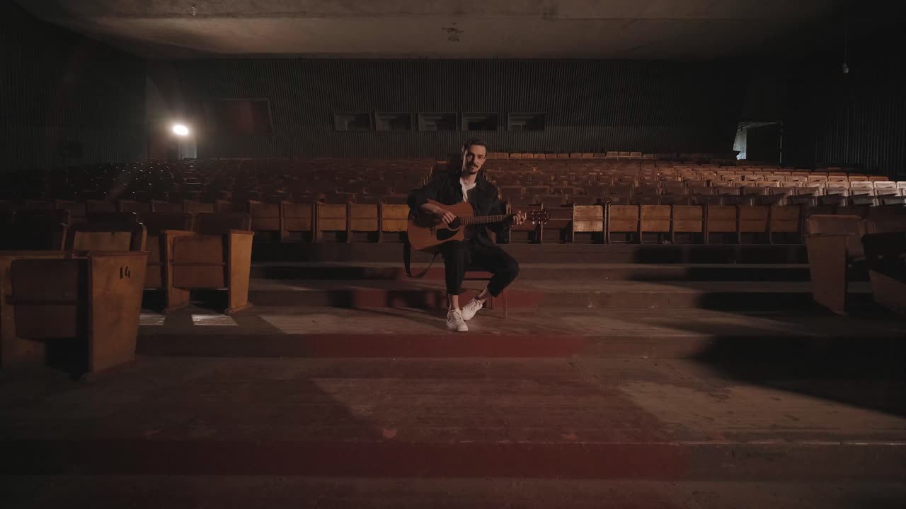 un chico guapo toca una guitarra acústica en un cine abandonado. el músico canta una canción y acompaña en la guitarra