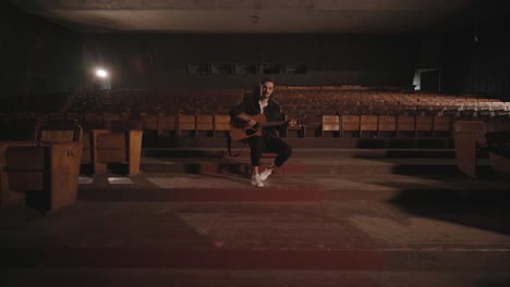 a handsome guy plays an acoustic guitar in an abandoned cinema. the musician sings a song and accompanies on the guitar