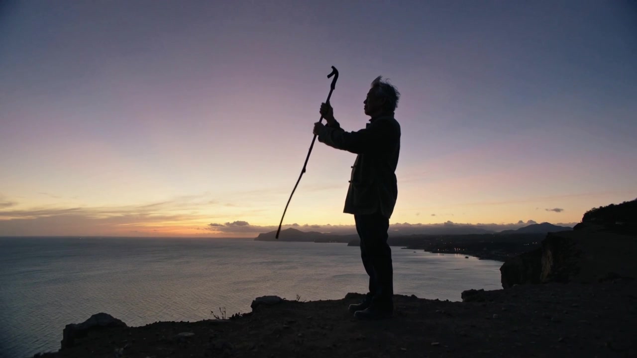 Escursionista anziano in piedi, in cappotto, che solleva il bastone su una scogliera mentre il cielo si oscura, esaminando l'orizzonte, con spazio per il testo