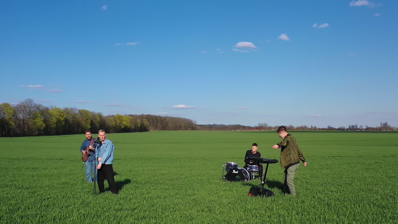 Young men perform music outdoors. Group of friends playing musical instruments and singing on green field background. Orbital view.