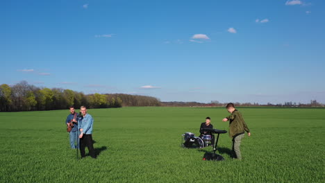 young men perform music outdoors. group of friends playing musical instruments and singing on green field background. orbital view.
