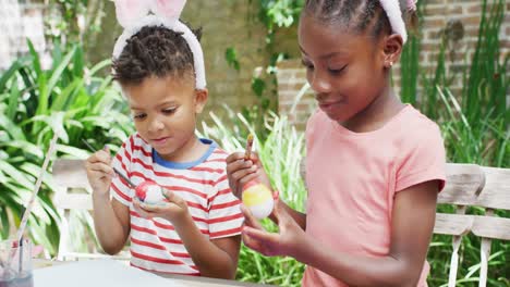 Happy-african-american-brother-and-sister-in-bunny-ears-painting-eggs-for-easter,-slow-motion