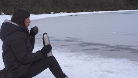 a woman drinking something hot in a very cold environment with snow and frozen lake