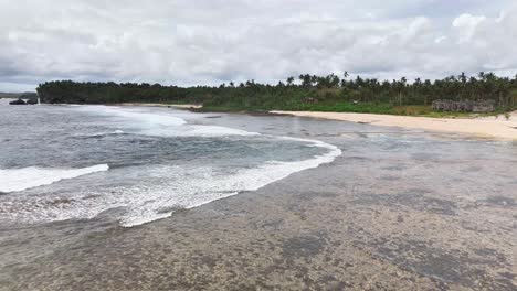 aerial view of magpupungko tidal pool in siargao islands, philippines with waves rolling over flat reef, white sand beach, rocky shoreline, and dense palm forest under overcast skies