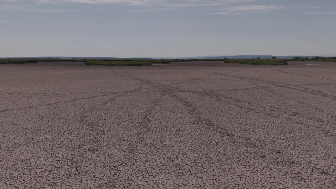 Um vasto leito de lago rachado se estende por um sapal seco sob um céu de verão claro, com ilhas isoladas de juncos visíveis no horizonte