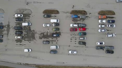 top down aerial footage of cars and vehicles parking in a parking lot during a cloudy day while people walk between to and from their cars. footage moves from right to left revealing more vehicles.