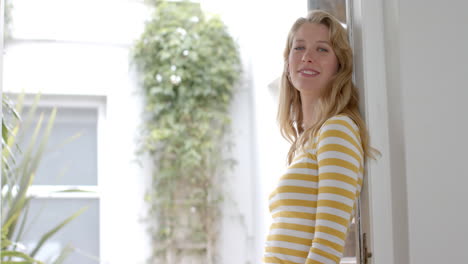 Portrait-of-happy-blonde-caucasian-woman-leaning-in-doorway-at-home,-copy-space,-slow-motion